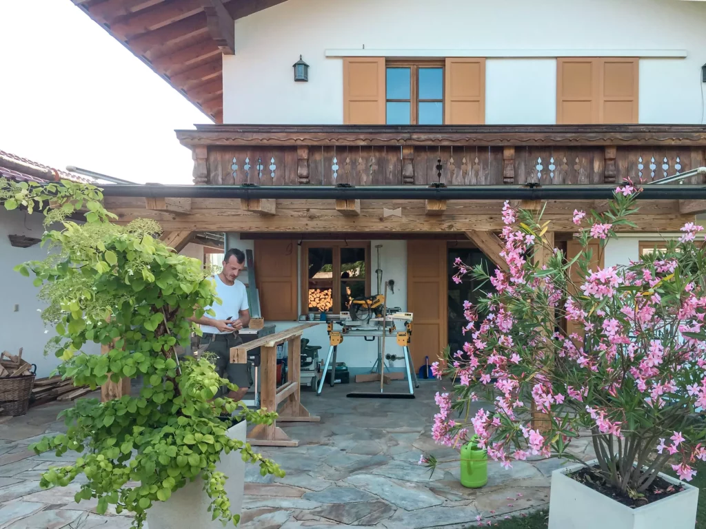 Tischler arbeitet an Altholzmöbeln vor traditionellem Haus mit Holzveranda und blühenden Pflanzen im Außenbereich