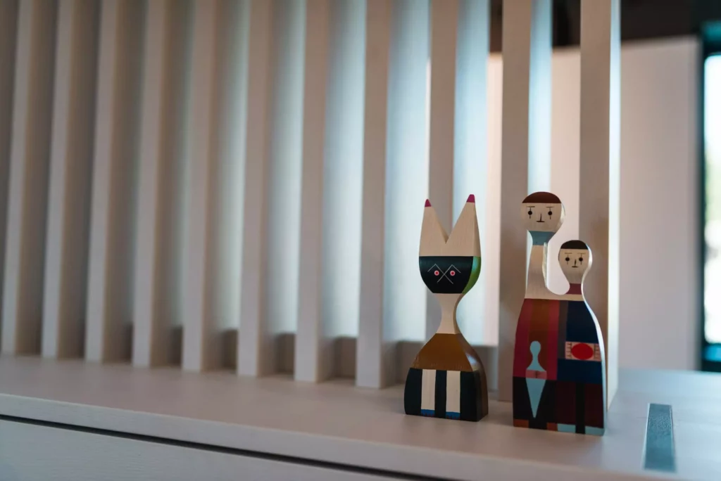 Zwei farbige, abstrakte Holzfiguren auf weißem Möbelstück vor vertikalen Holzlamellen im Showroom des Möbelstudios M-Studio