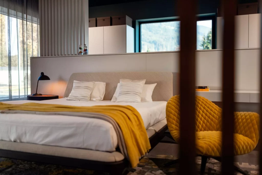 Modernes Schlafzimmer im Möbelstudio mit gepolstertem Bett, gelbem Design-Sessel, Tagesdecke und großem Fenster im Showroom