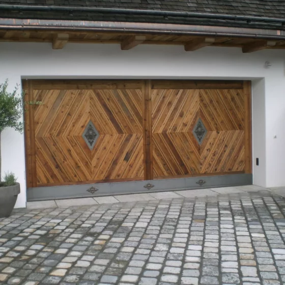 Garagentor aus Holz mit diagonalem Muster und Metallverzierungen, Pflanze in grauem Topf davor, Außenbereich, Weitgasser