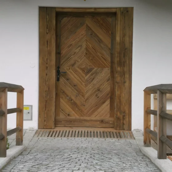 Eingangstür aus Holz mit Fischgrätmuster und Holzgeländer an gepflastertem Weg vor weißer Hauswand, Tischlerei Weitgasser