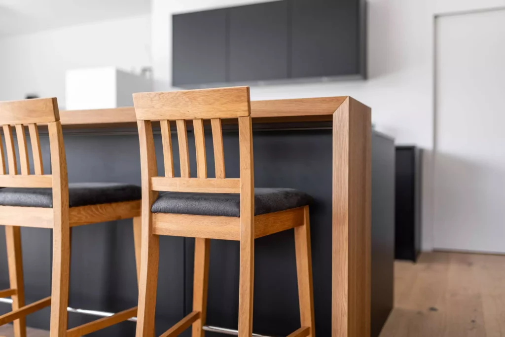 Moderne Tischlereiküche mit Holztheke und zwei Holzstühlen mit schwarzen Sitzpolstern in minimalistischem, Tischlerküche
