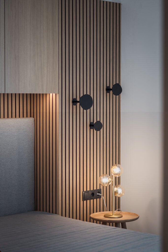 Moderner Schlafzimmerbereich mit grauem Polsterbett, Holzpaneelwand mit schwarzen Haken und Design-Tischleuchte, Einrichtung