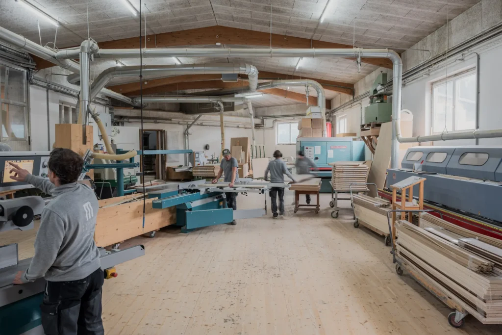 Holzwerkstatt mit drei Arbeitern, modernen Maschinen und Holzplatten in einem hellen Raum mit sichtbaren Lüftungsrohren