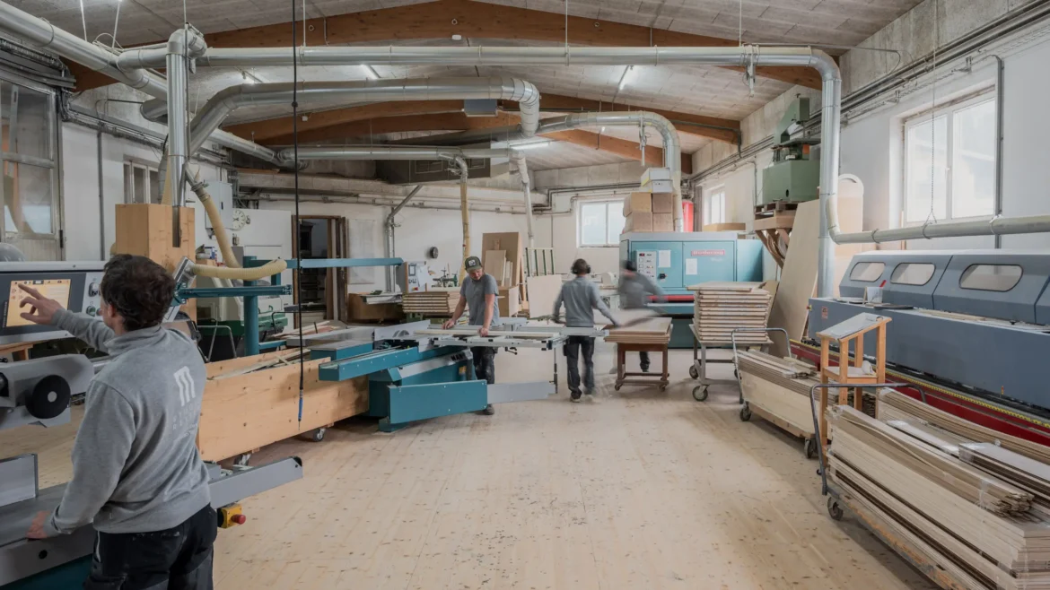 Holzwerkstatt mit drei Arbeitern, modernen Maschinen und Holzplatten in einem hellen Raum mit sichtbaren Lüftungsrohren