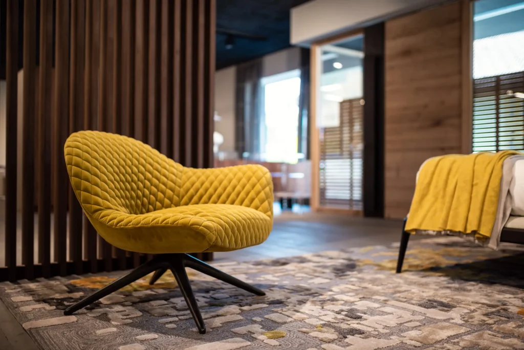 Gelber, gepolsterter Design-Sessel auf gemustertem Teppich vor Holzlamellen in modernem Möbelstudio